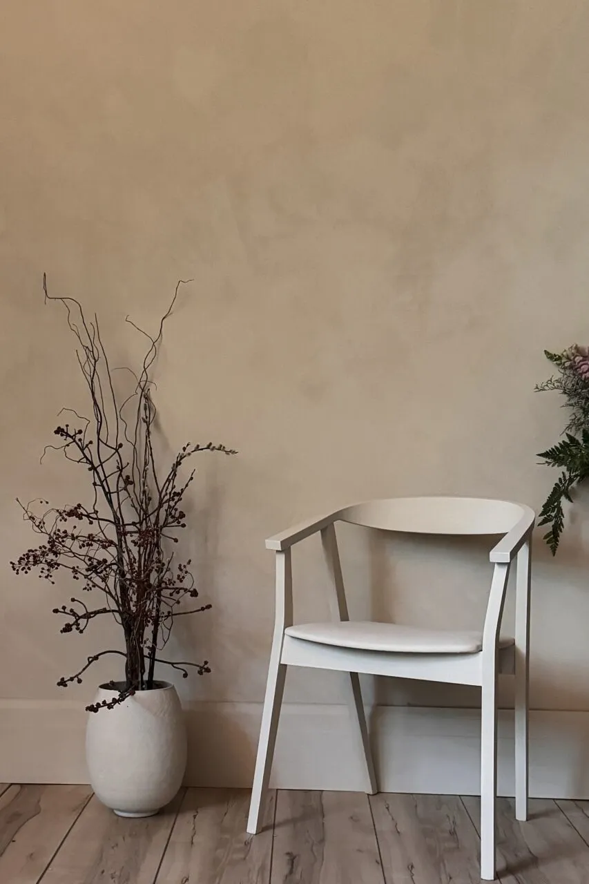 Wall in warm sand limewash, its soft brush movement behind a pale wood chair and a stoneware vase of dried branches.